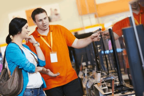 A man in an orange shirt with a lanyard pointing to monitors in a store with a woman listening with her head resting on her hand